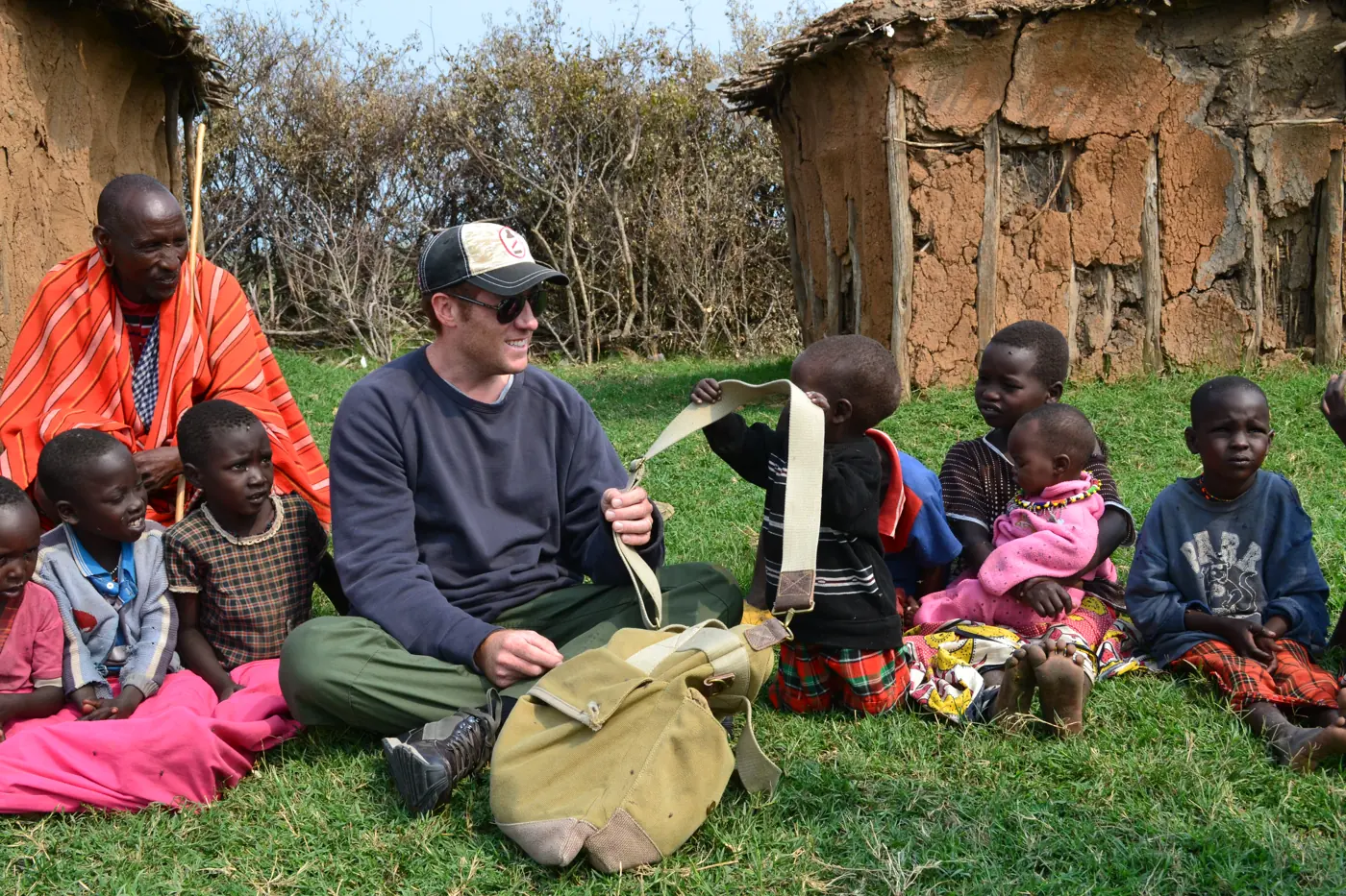 A man sitting in the grass surrounded by smiling Maasai children in front of mud houses